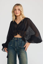 A woman modeling a black sheer long-sleeve crop top with ruffled sleeves and dark blue wide-leg denim jeans, standing against a light gray studio background.
